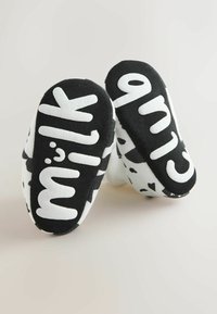 Zapatillas negras y blancas sin cordones con el texto grande "milk" y "clue" en una superficie lisa, con un diseño de estampado de vaca manchada en los laterales.