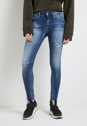 Osoba nosící vybledlé modré úzké džíny s mírným opotřebením na stehnech a kolenou, černé slip-on tenisky s bílými podrážkami a tmavý svetr.