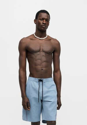 Homme à la peau foncée et aux cheveux courts porte un short de bain bleu clair à cordon et un collier de perles blanches, debout devant un fond blanc uni.