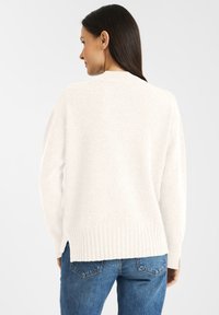Biały sweter dziany z prążkowanym dołem, długimi rękawami i bocznymi rozcięciami, noszony na niebieskie dżinsy. Miękka tekstura, casualowy design.