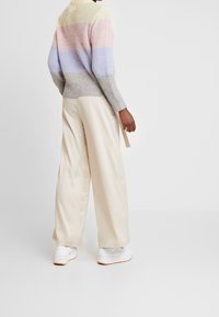 Gebreide trui met pastel horizontale strepen in roze, lila en grijs, gecombineerd met crèmekleurige wijde broek en witte sneakers. Achteraanzicht.