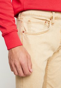 Beige denim jeans with copper rivets, paired with a bright red sweatshirt. The jeans have a smooth texture and traditional five-pocket design.