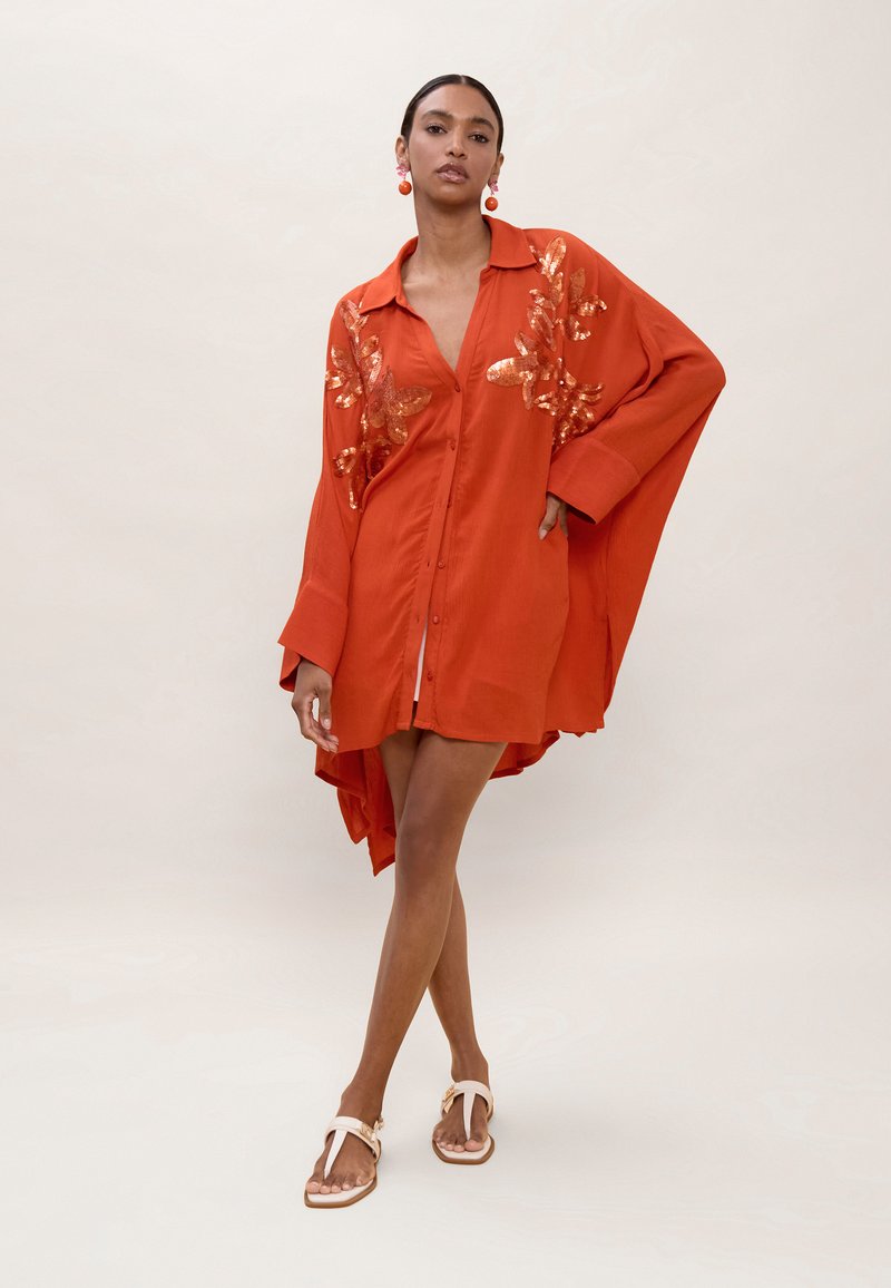 Nainen seisoo jalat ristissä yllään ylisuurikokoinen oranssi paitamekko, jossa kiiltävät kukkakuviot, sekä valkoiset sandaalit, yksiväristä taustaa vasten.