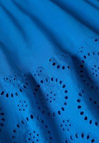 Tissu bleu vif avec des motifs circulaires brodés et des œillets, montrant des plis et des détails de texture.