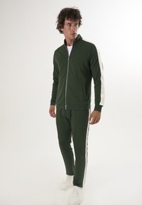 Grüner Zip-Jacke mit weißen Seitenstreifen, kombiniert mit passenden schmal geschnittenen Hosen. Beide Teile sind aus einem glatten, dehnbaren Stoff gefertigt. Weiße Sneaker.