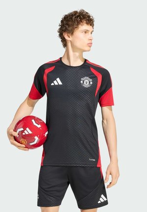 Giovane uomo con la divisa nera e rossa del Manchester United che tiene una palla da calcio rossa, guardando verso destra.