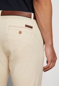 Pantalones beige claros, hechos de una tela suave, con un cinturón de cuero marrón, bolsillos laterales y un pequeño parche con el logo en la parte trasera.