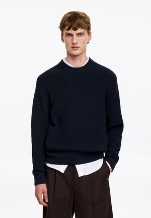 Jeune homme aux cheveux courts châtain clair portant un pull en maille bleu marine par-dessus une chemise blanche et un pantalon sombre, debout devant un fond blanc uni.