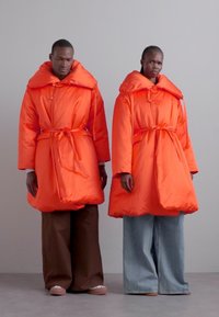 Δύο άτομα φορούν μεγάλα, φουσκωτά μπουφάν σε έντονο πορτοκαλί χρώμα, με κουκούλες και ζώνες στη μέση, σε συνδυασμό με φαρδιά παντελόνια από καφέ και μπλε τζιν.