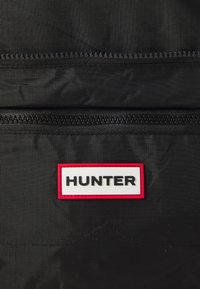 Fekete nejlon hátizsák, amely két cipzárral, textúrált anyaggal rendelkezik, és egy fehér címkével, piros kerettel, amelyen vastag fekete betűkkel "HUNTER" felirat található.