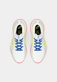 Des chaussures de sport blanches avec des lacets bleus, des accents jaunes et une semelle rouge. Elles présentent une semelle intérieure rembourrée et une tige en mesh pour une meilleure respirabilité.