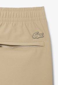 Pantalon beige avec taille élastique, poche zippée au dos et petit logo crocodile brodé sur le côté droit.