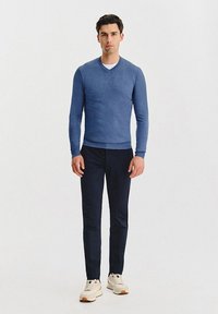 Niebieski sweter w serek z miękkiego dzianiny, zestawiony z ciemnymi spodniami i białymi sneakersami. Strój ma opływowy, dopasowany wygląd.