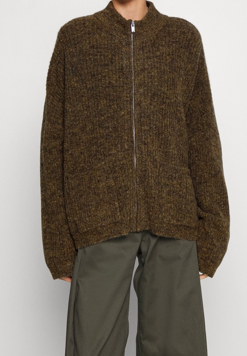 Person, der eine braune, strukturierte Zip-Up-Strickjacke mit zwei vorderen Taschen über lockeren olivgrünen Hosen trägt, die bis zur Mitte des Oberschenkels geschnitten sind.