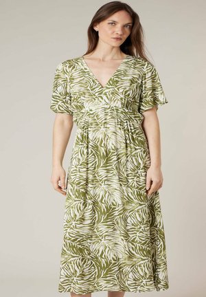 Femme portant une robe midi à manches courtes et col en V, à motif de feuilles vertes et blanches, debout devant un fond beige uni.