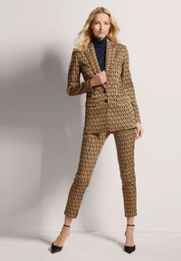 Bruin gestippeld pak met zwarte abstracte ontwerpen, aansluitende vorm, contrasterende marineblauwe coltrui en zwarte stilettohakken. Op maat gemaakt jasje en broek.