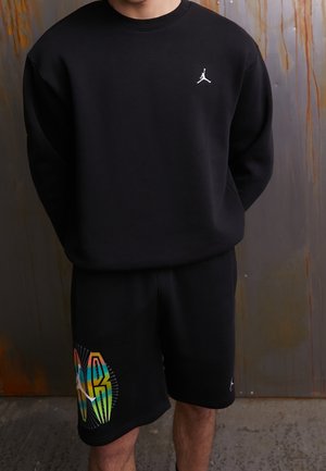 Osoba ubrana w czarną bluzkę z długim rękawem z białym logo Jumpman oraz czarne krótkie spodenki z kolorowym napisem "JORDAN" i logo Jumpman.