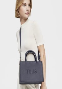 Bolso de piel color marino con dos asas superiores y una correa de hombro ajustable. Presenta el logotipo grabado "TOUS" y una superficie texturizada.