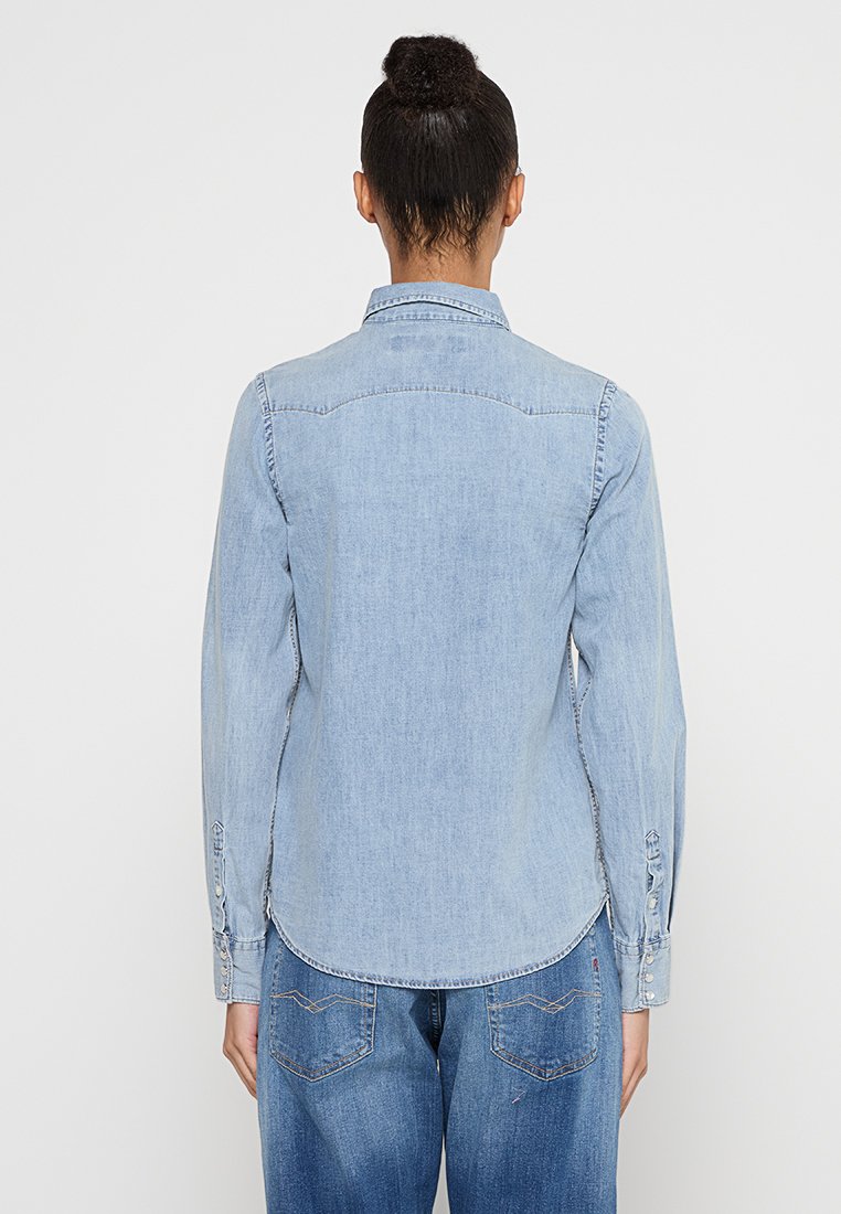 Persona con el cabello oscuro recogido en un moño, vistiendo una camisa de mezclilla azul clara y jeans azules, vista desde atrás contra un fondo sencillo.