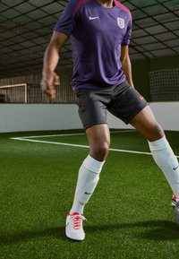 Manlig fotbollsspelare i engelsk tröja och fotbollsskor, som kontrollerar bollen på inomhusgräs nära målstolpen.