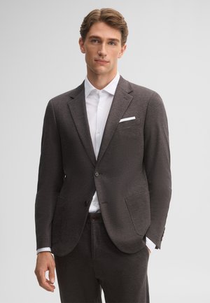 Jonge man met bruin haar draagt een donkergrijs colbert en broek over een wit overhemd, staand tegen een lichtgrijze achtergrond.