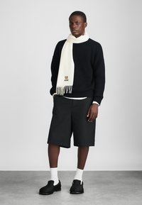 Μαύρο πλεκτό πουλόβερ με ραβδώσεις, κρεμ φουλάρι με φ fringe και λογότυπο, μαύρες βερμούδες και υφασμάτινα μαύρα παπούτσια χωρίς κορδόνια. Επίπεδη εμφάνιση με λευκές λεπτομέρειες.