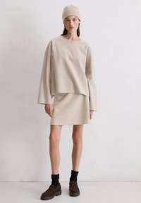 Beżowy sweter z dzianiny o szerokich rękawach, noszony na pasującą spódnicę. Uzupełniony ciemnymi skarpetkami i brązowymi butami wsuwanymi.