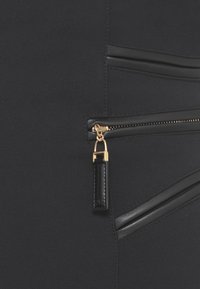 Tessuto nero con una superficie liscia che presenta una tasca con zip inclinata. La zip è dorata e ha un cursore rettangolare in pelle nera.