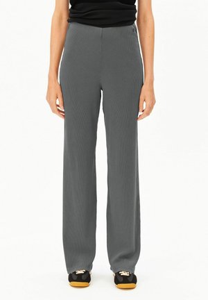 Frau trägt locker sitzende, hochtaillierte graue Hose und schwarze Sneakers mit beigen Schnürsenkeln, steht vor einem schlichten weißen Hintergrund.