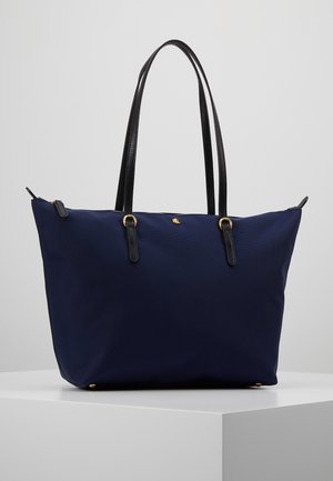 Bolsa tote azul marino con asas de cuero negro, herrajes dorados y un pequeño logo dorado en el frente, colocada sobre un pedestal blanco.