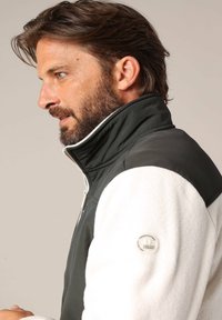 Profil d'un homme avec une barbe portant une veste en polaire zippée noire et blanche avec un patch logo sur la manche, sur fond beige.