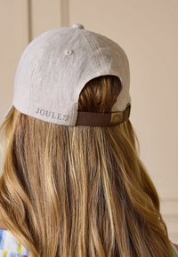 Beežist linast pesapalli nokamüts reguleeritava pruunist rihmaga, kantud üle pika blondi juukse, tagumine vaade, brändinimi "Joules" mütsil.