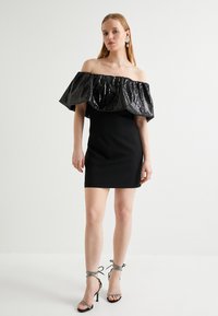 Robe noire avec une silhouette cintrée, ornée d'un vol lumineux de sequins en volants dénudés sur les épaules. Accompagnée de talons hauts à lanières.
