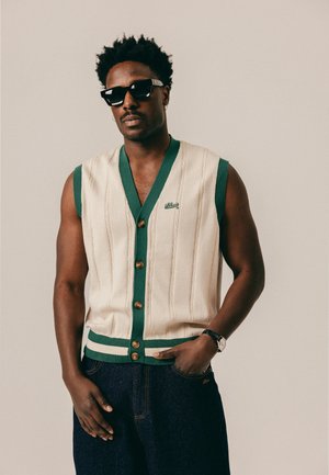 Man wearing black sunglasses, sleeveless cream sweater vest with green trim and buttons, dark jeans, and a wristwatch, standing against plain background.