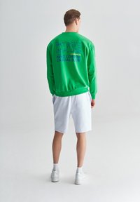 Zielona bluza z grafiką na plecach, zestawiona z białymi szortami. Ubranie ma luźny krój i ribbowe mankiety. Białe sneakersy dopełniają całość.