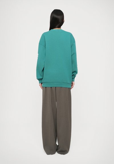 Felpa turchese con spalle cadenti e polsini a costine, abbinata a pantaloni grigi larghi. Texture liscia e vestibilità oversized.