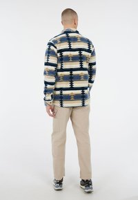 Man in beige cargobroek, zwart-witte sneakers en een blauw, beige en wit gestreepte lange mouwen shirt, met zijn rug naar de camera op een witte achtergrond.