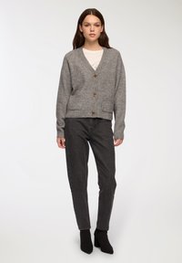 Szary dziany kardigan z dekoltem w serek, pięcioma brązowymi guzikami i dwiema przednimi kieszeniami, zestawiony z ciemnymi dżinsami i czarnymi botkami za kostkę.