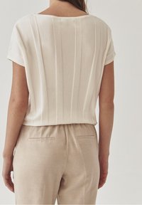 Top avorio a maniche corte con pieghe verticali, abbinato a pantaloni beige con tasche posteriori piatte e cintura elastica.