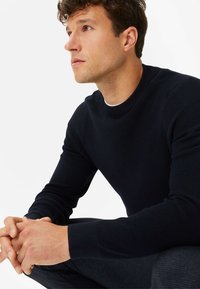 Dunkelblauer Crewneck-Pullover aus Rippstrick mit langen Ärmeln und taillierter Silhouette. Das Modell sitzt mit gefalteten Händen.