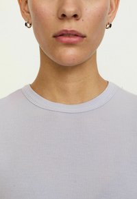 Svijetloljubičasti rebrasti top s okruglim izrezom. Jednostavan dizajn naglašen malim kružnim naušnicama. Glatka, mekana tekstura tkanine.