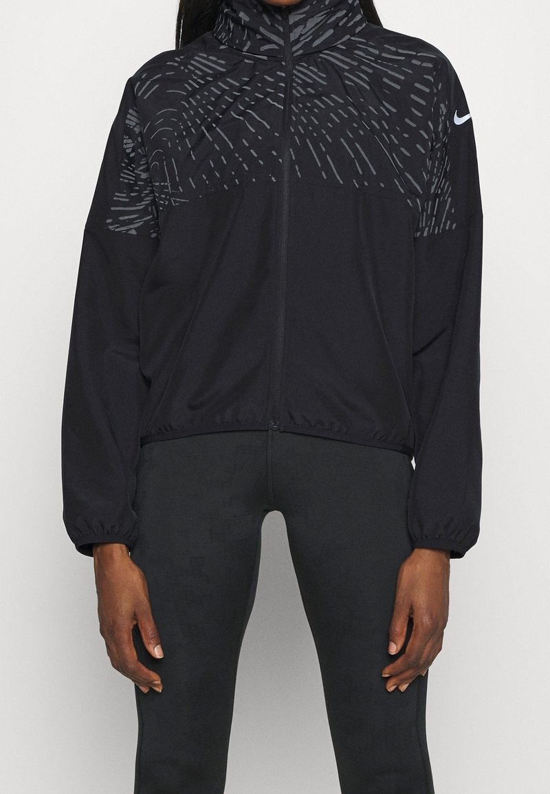 Veste de sport noire zippée intégralement avec un motif gris subtil sur les épaules et les manches, portée avec un pantalon noir ajusté, logo Nike blanc sur la manche gauche.