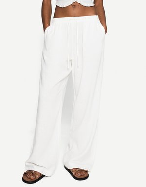 Model wears white, loose-fitting pants with a drawstring waist, paired with a cropped top and brown sandals, hands in pockets.