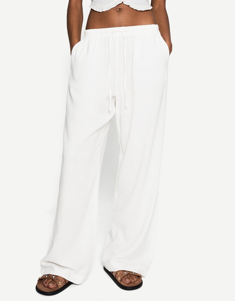 Le modèle porte un pantalon blanc, ample, avec une taille à cordon, associé à un top court et des sandales brunes, les mains dans les poches.