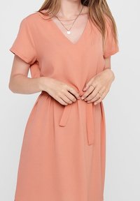 Persikofärgad klänning med V-hals och korta ärmar, smal midja och avtagbart knytbälte. Slät textur och flytande siluett. Enkel design.