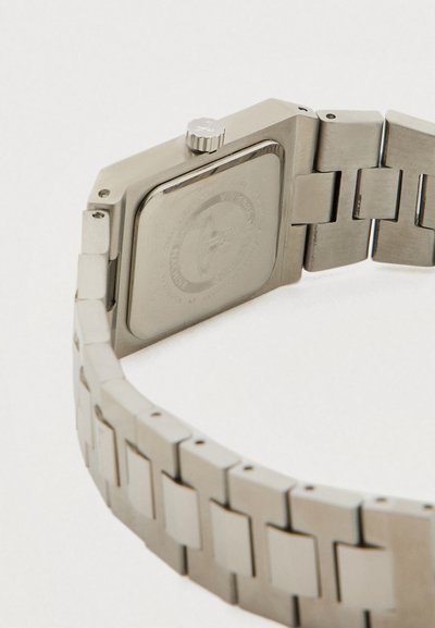 Βραχιόλι ρολογιού χειρός από ανοξείδωτο ατσάλι με μεταλλικούς κρίκους και τετράγωνη θήκη ρολογιού που φαίνεται από πίσω, εμφανίζοντας την κορώνα και το χαραγμένο λογότυπο.