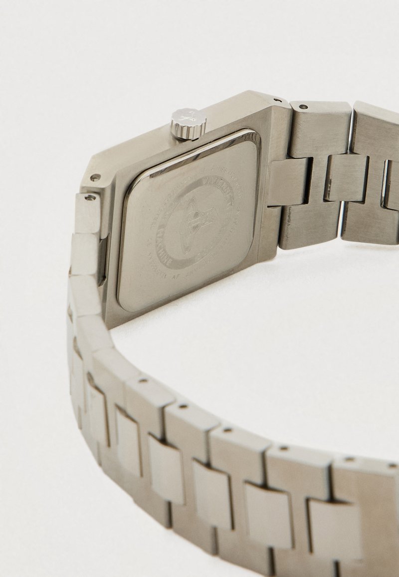 Roestvrijstalen horlogeband met metalen schakels en een vierkante horlogekast, gezien vanaf de achterkant, met zichtbaar de kroon en gegraveerd logo.