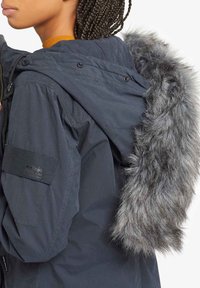 Navy blue parka jacket with a faux fur hood trim. Features metal snap buttons and a textured fabric. Adjustable drawstring at the hood.