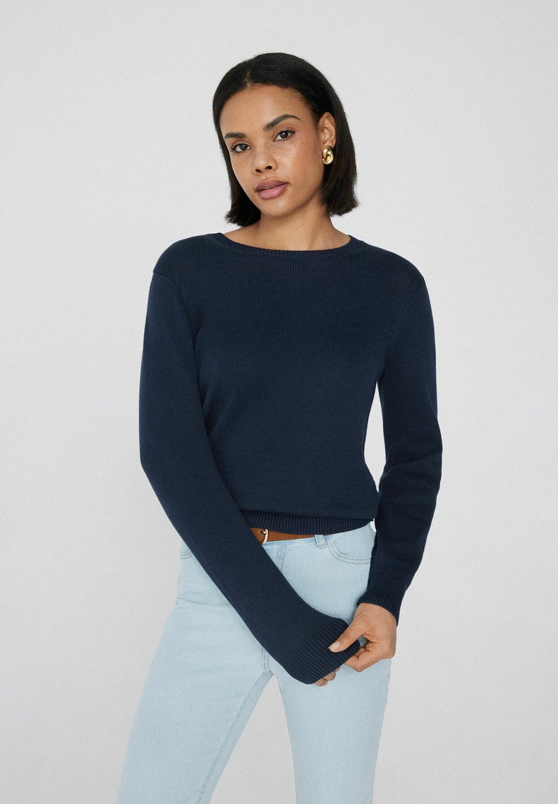 Marineblauer Rippstrickpullover mit langen Ärmeln, horizontalem Ausschnitt und umgeschlagenem Saum, kombiniert mit hellblauen Jeans. Glatte Textur.