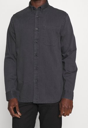 Homme portant une chemise à manches longues gris foncé boutonnée avec une poche poitrine avant et un pantalon noir sur un fond blanc uni.